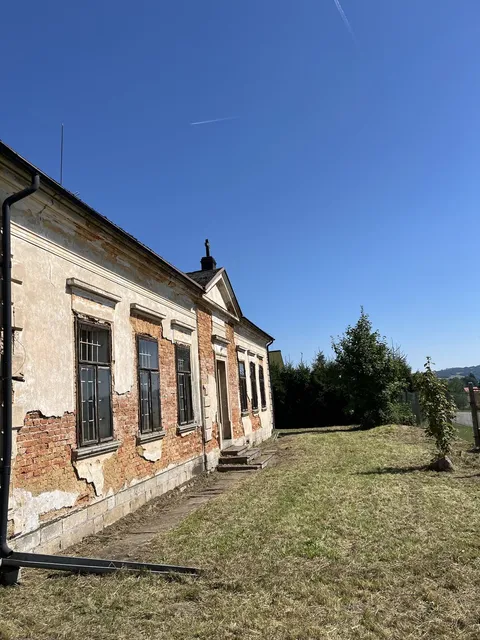 Dlouhá fasáda starého statku s popraskanou omítkou, podél zdi čerstvě posekaná tráva